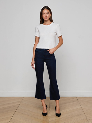 A woman stands indoors against a plain white wall, wearing the Stormy Cropped Cotton Tee in white, dark blue cropped flare jeans, and black heels. She has her left hand in her pocket and looks at the camera.