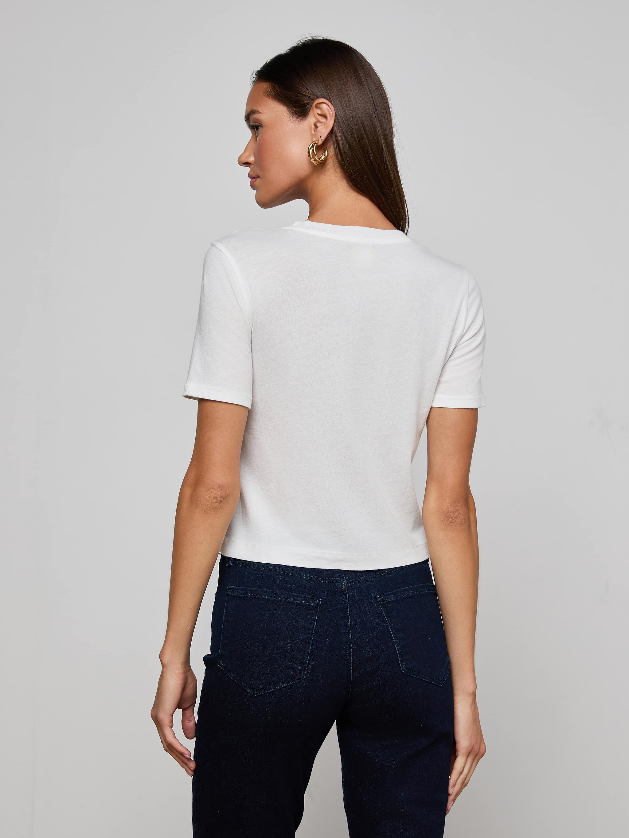 A person with long blonde hair is shown from behind, wearing the Stormy Cropped Cotton Tee in plain white and blue jeans against a light background.