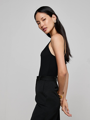A woman with long black hair stands in profile against a light gray background, wearing the L'AGENCE Naseem Ruched Bodysuit with adjustable straps, black pants, gold earrings, and a bracelet, her hands kept elegantly behind her back.
