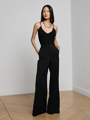 A woman with long straight hair stands in a minimalist room wearing the L'AGENCE Naseem Ruched Bodysuit and high-waisted, wide-leg black pants, accessorized with a gold necklace and bangles, hands casually placed in her pockets.