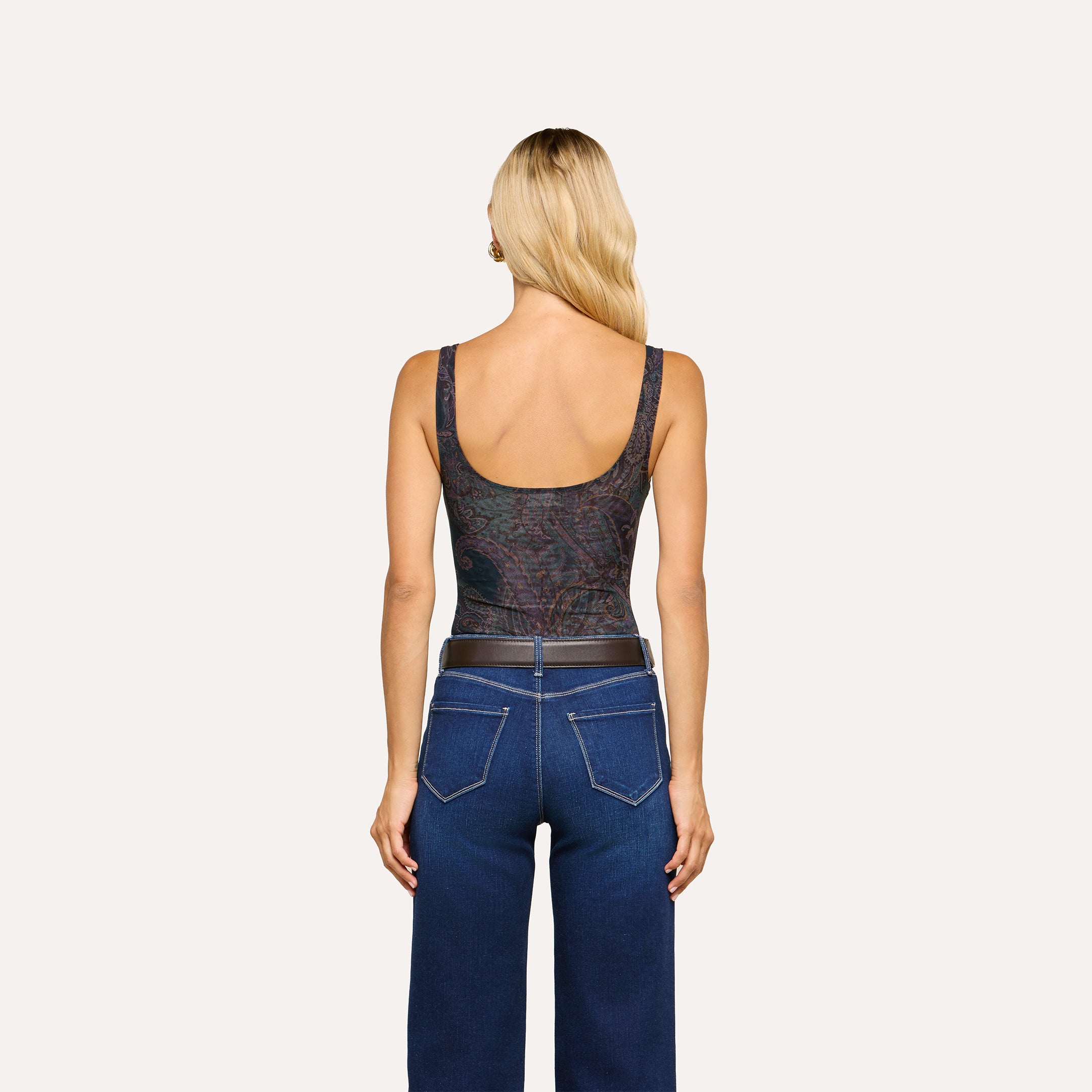 A woman with long blonde hair stands facing away, wearing the sleeveless midnight blue Kalea Mesh Bodysuit featuring a paisley pattern, paired with high-waisted blue jeans with visible stitching and a black belt, set against a plain light background.