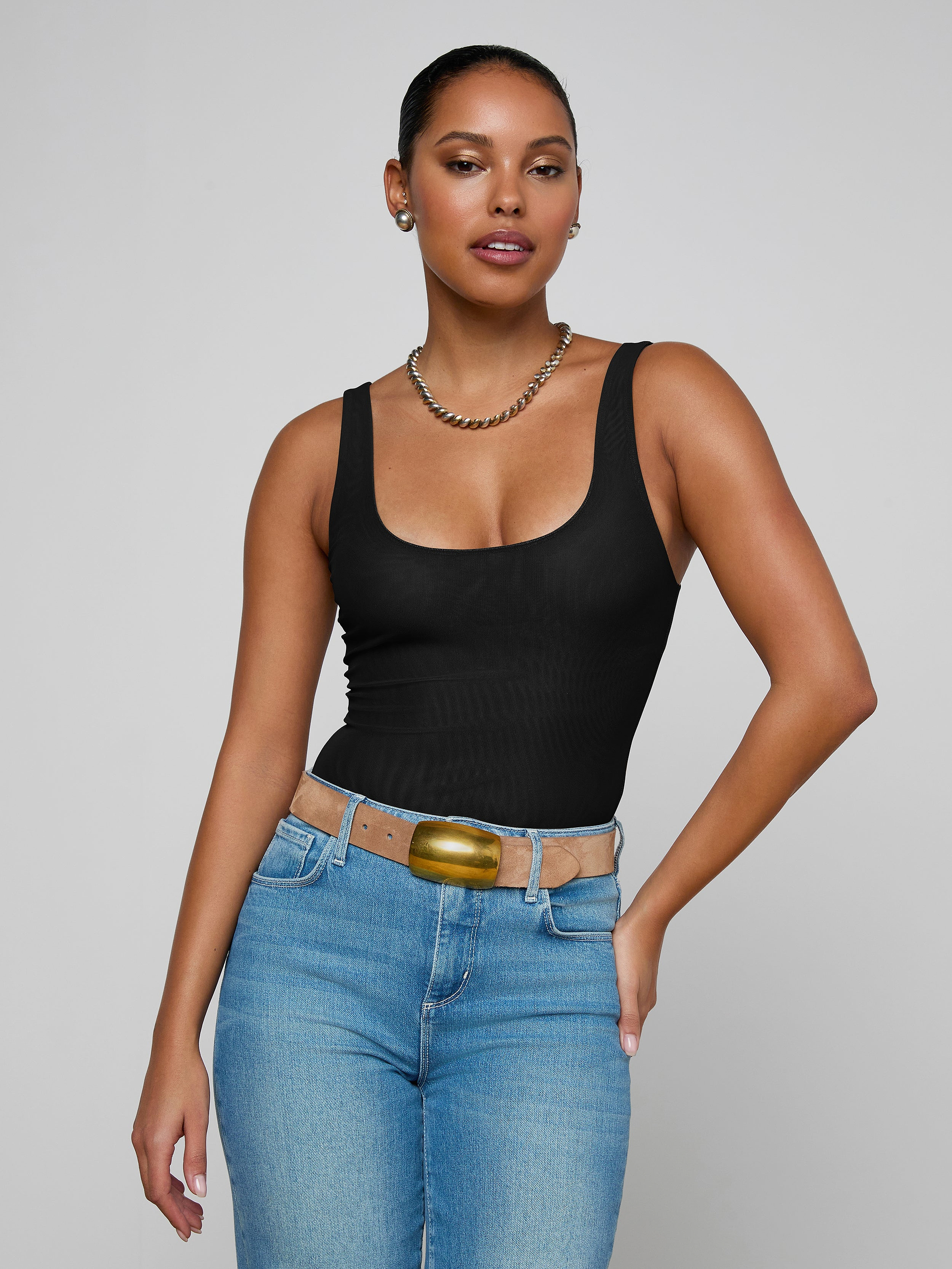A woman with dark hair pulled back is wearing the Kalea Mesh Bodysuit, styled with a gold necklace, tan belt, and blue jeans. She stands against a plain light background with one hand on her hip.