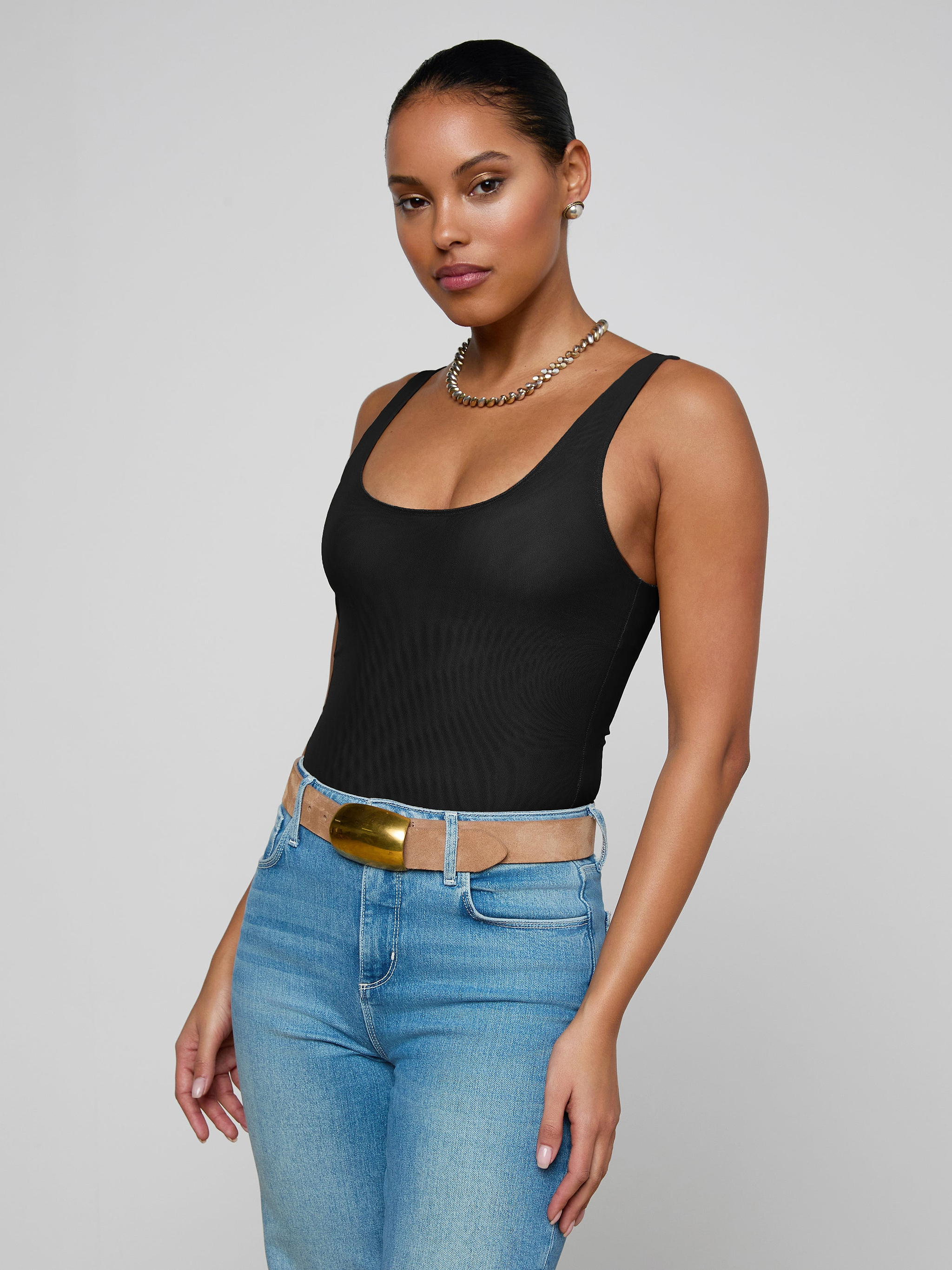 A woman stands against a plain background, wearing the Kalea Mesh Bodysuit, blue jeans with a tan belt, and a gold chain necklace. Her hair is pulled back as she looks confidently at the camera.