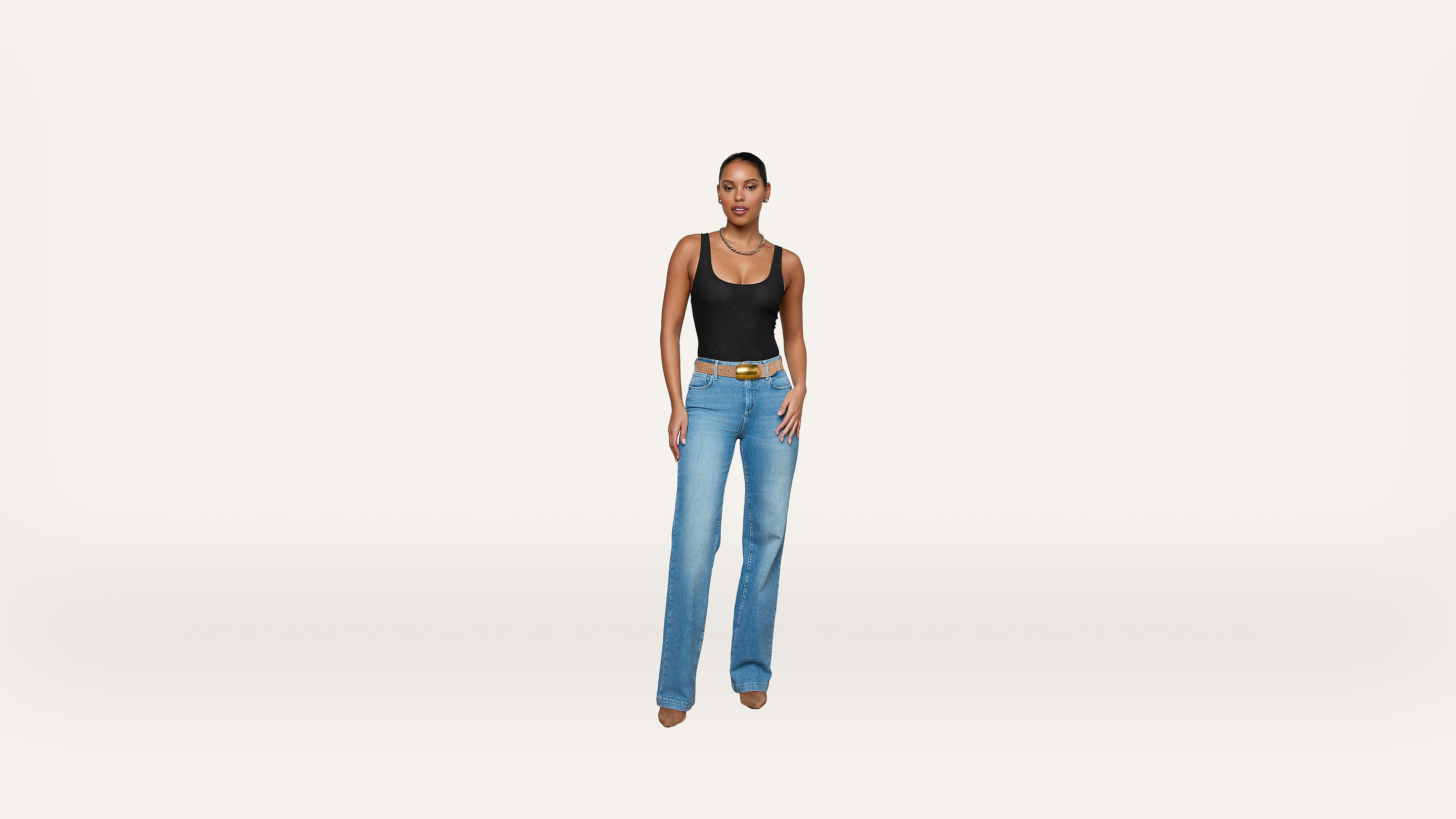 A woman stands against a plain white background, wearing a black tank top, blue jeans, a brown belt, and beige shoes.