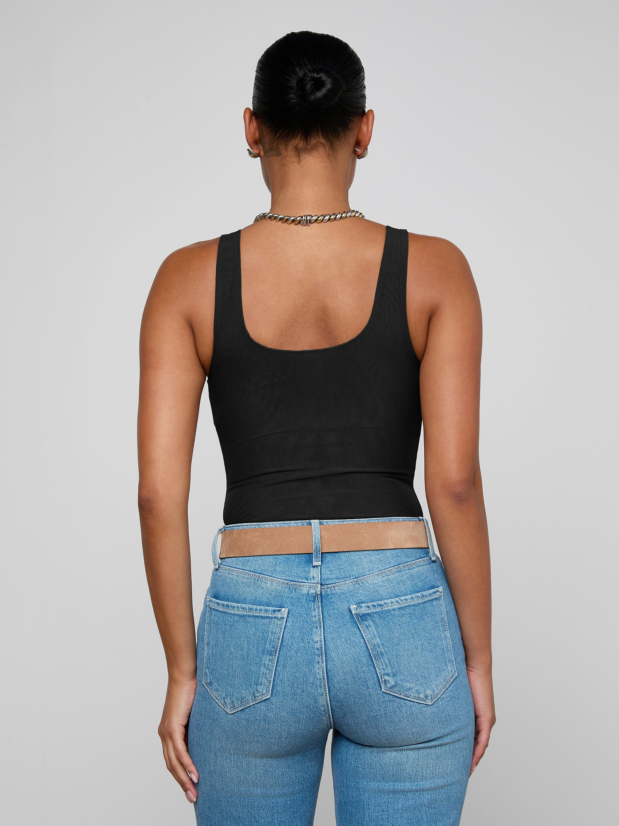 A person with hair in a bun, wearing the Kalea Mesh Bodysuit, blue jeans, and a chain necklace, is seen from the back against a plain light background.