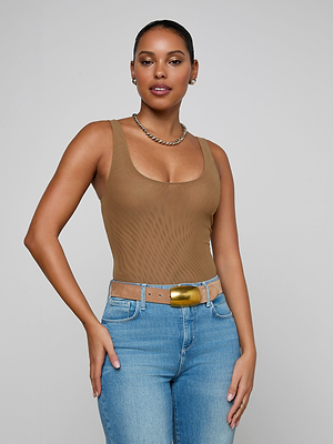 A woman poses confidently against a light gray background wearing the sleeveless brown Kalea Mesh Bodysuit, gold necklace, and blue jeans with a tan belt.