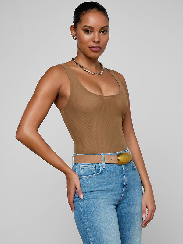 A woman with long brown hair is wearing the Kalea Mesh Bodysuit in a lightweight tan fabric, paired with blue jeans and a belt. She stands in profile against a plain light background, accessorized with gold earrings and sporting a neutral expression.