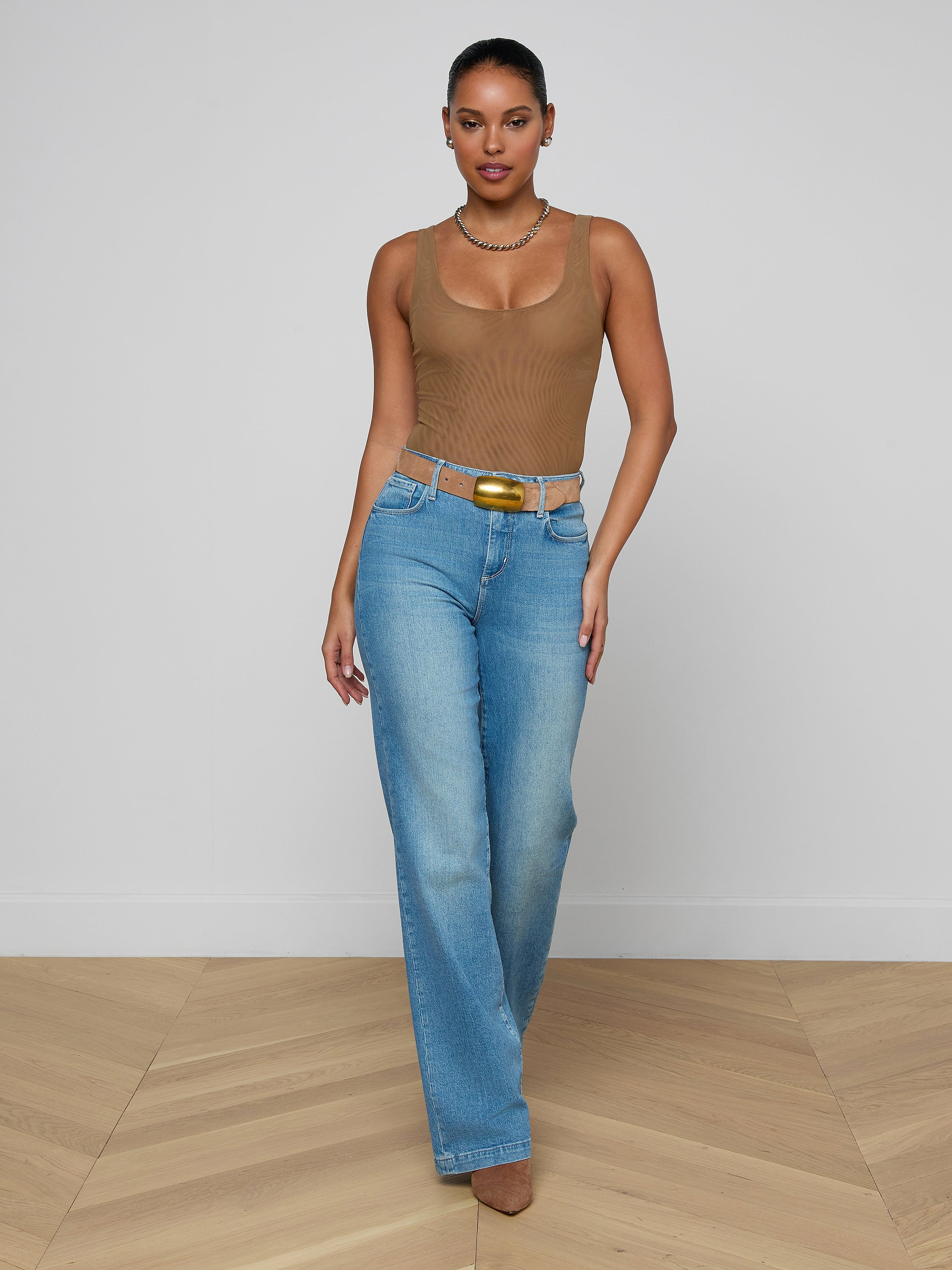 A woman stands against a plain white background wearing sunglasses, blue jeans with a belt, heeled sandals, and the Kalea Mesh Bodysuit in tan. She faces forward with her arms relaxed at her sides.