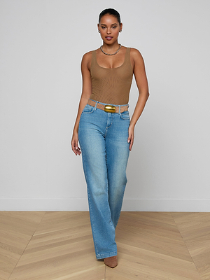 A woman stands in a neutral-toned room wearing the Kalea Mesh Bodysuit, light blue high-waisted jeans with a belt, brown shoes, and a necklace. Her hair is pulled back as she confidently looks at the camera.