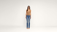 A woman wearing sunglasses, a tan tank top, blue jeans, and sandals stands facing forward against a plain white background.
