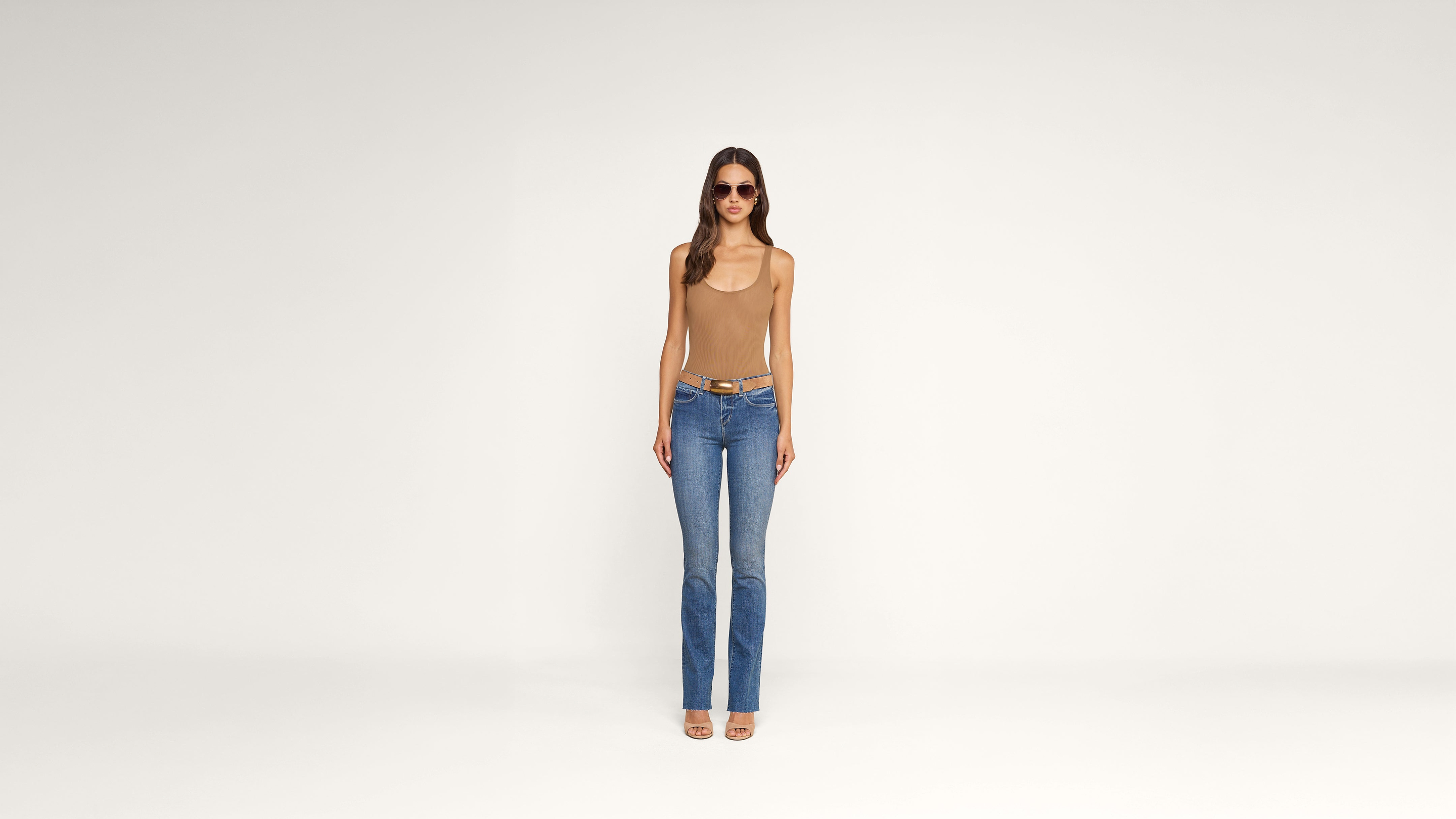 A woman wearing sunglasses, a tan tank top, blue jeans, and sandals stands facing forward against a plain white background.