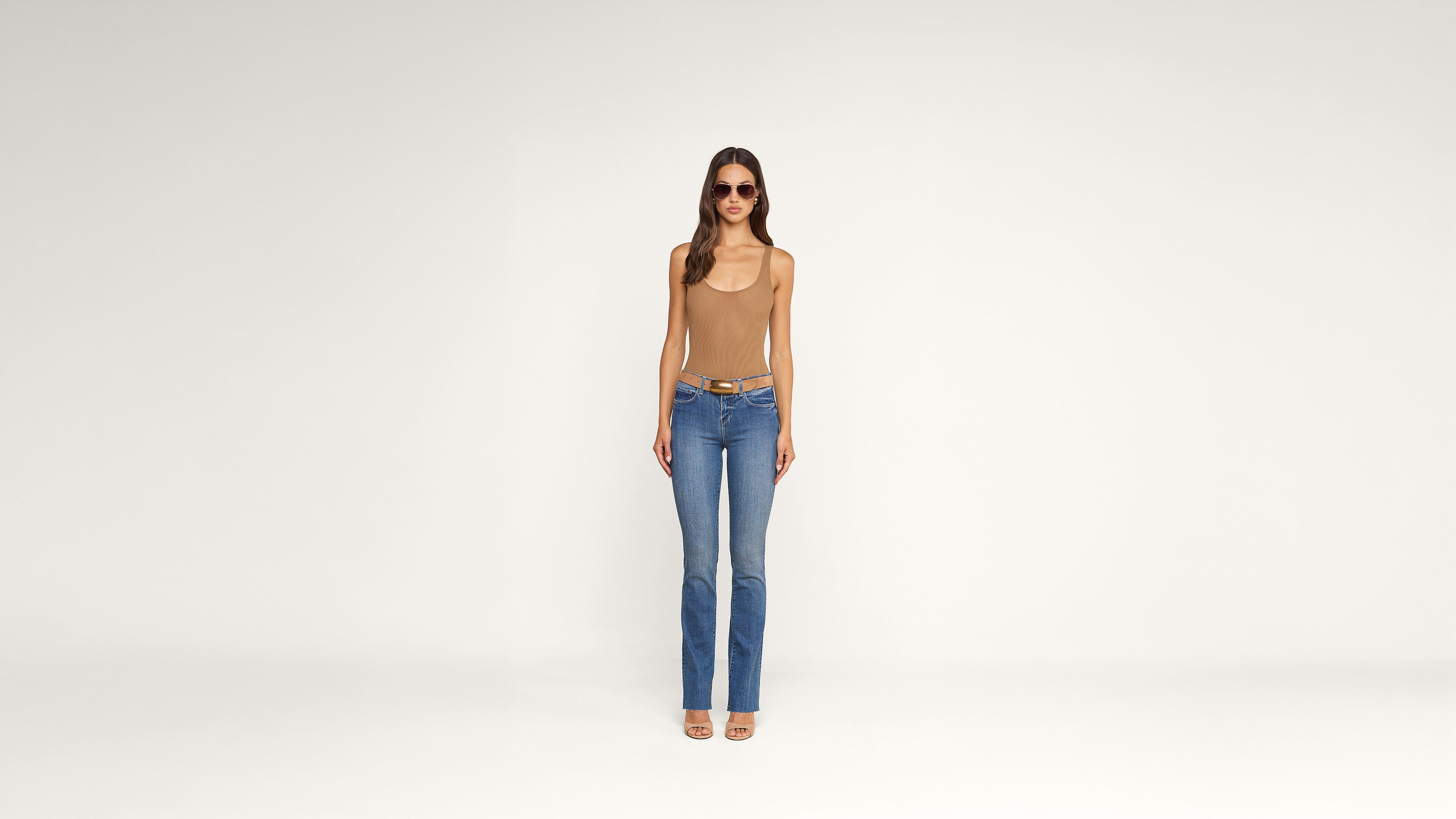 A woman wearing sunglasses, a tan tank top, blue jeans, and sandals stands facing forward against a plain white background.