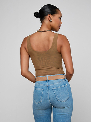 A woman with her hair in a bun wears the Kalea Mesh Bodysuit, a brown sheer stretch mesh sleeveless top, paired with light blue high-waisted jeans, and accessorized with a chain necklace and small hoop earrings.