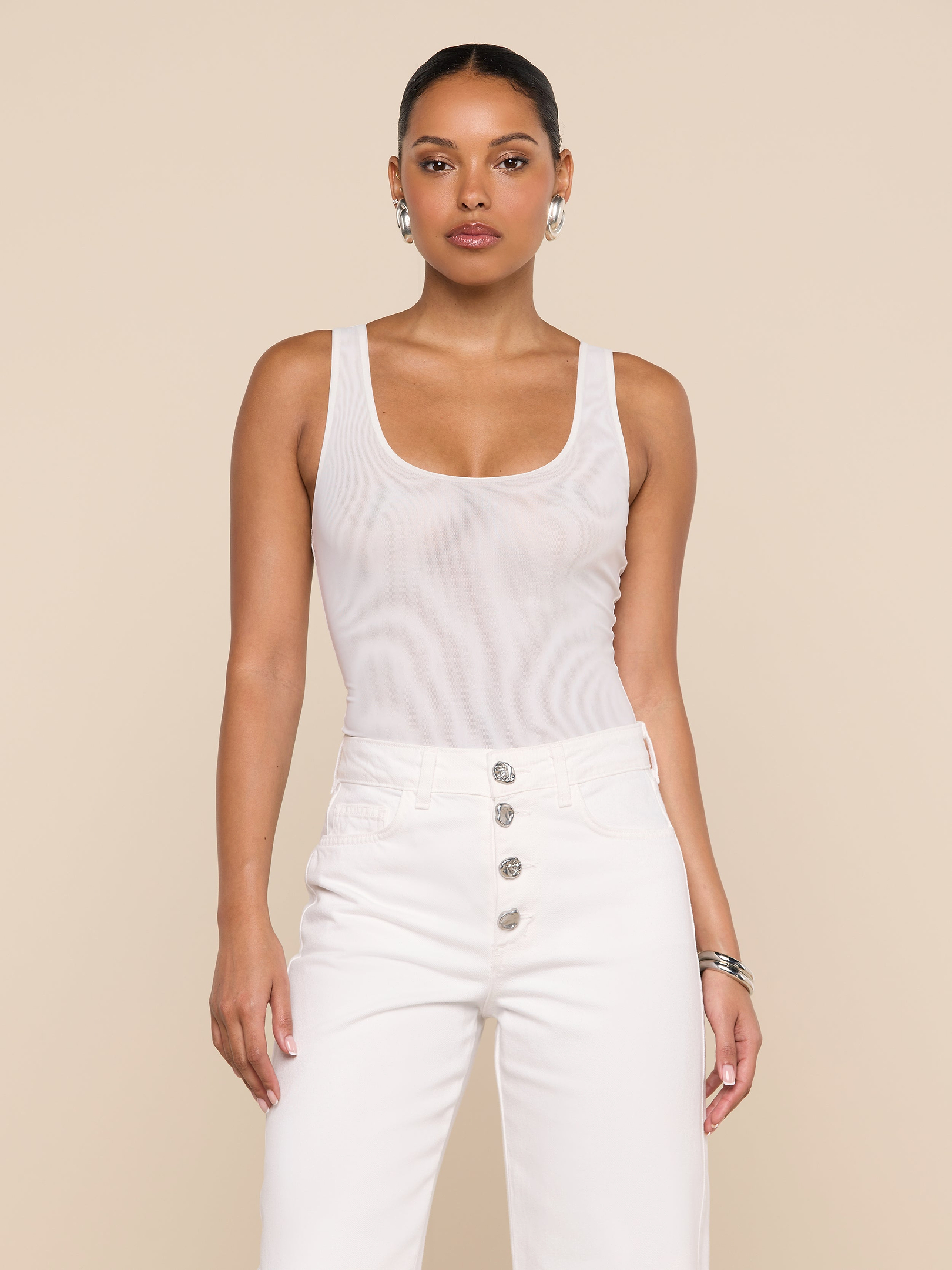A woman stands facing forward against a plain light background, wearing the Kalea Mesh Bodysuit—a sleeveless white mesh piece—paired with high-waisted white pants featuring silver buttons.