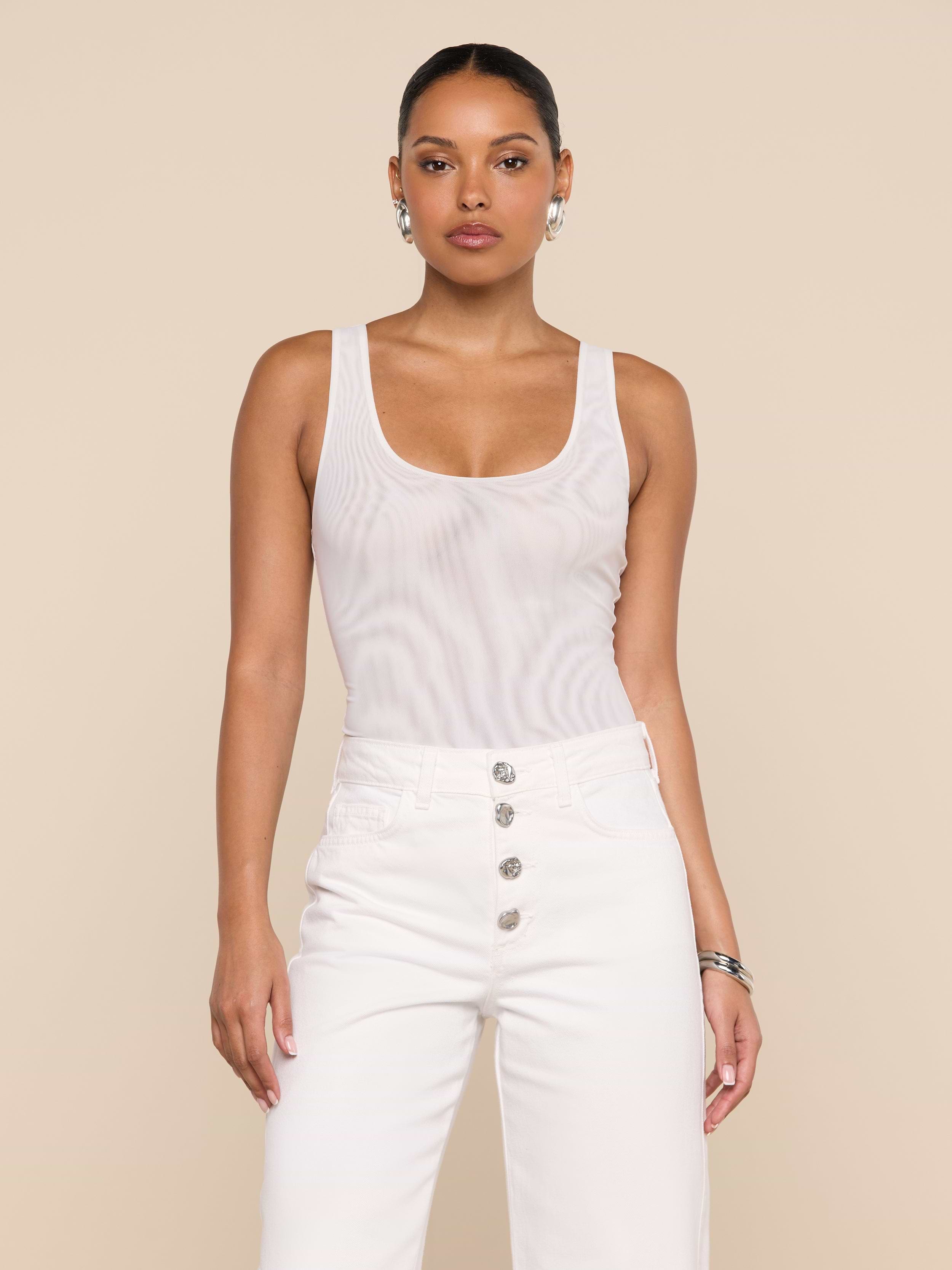 A woman stands facing forward against a plain light background, wearing the Kalea Mesh Bodysuit—a sleeveless white mesh piece—paired with high-waisted white pants featuring silver buttons.