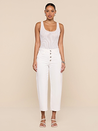 A woman stands facing forward, styled in the Kalea Mesh Bodysuit beneath high-waisted white pants with buttons and white sandals, set against a plain white background.