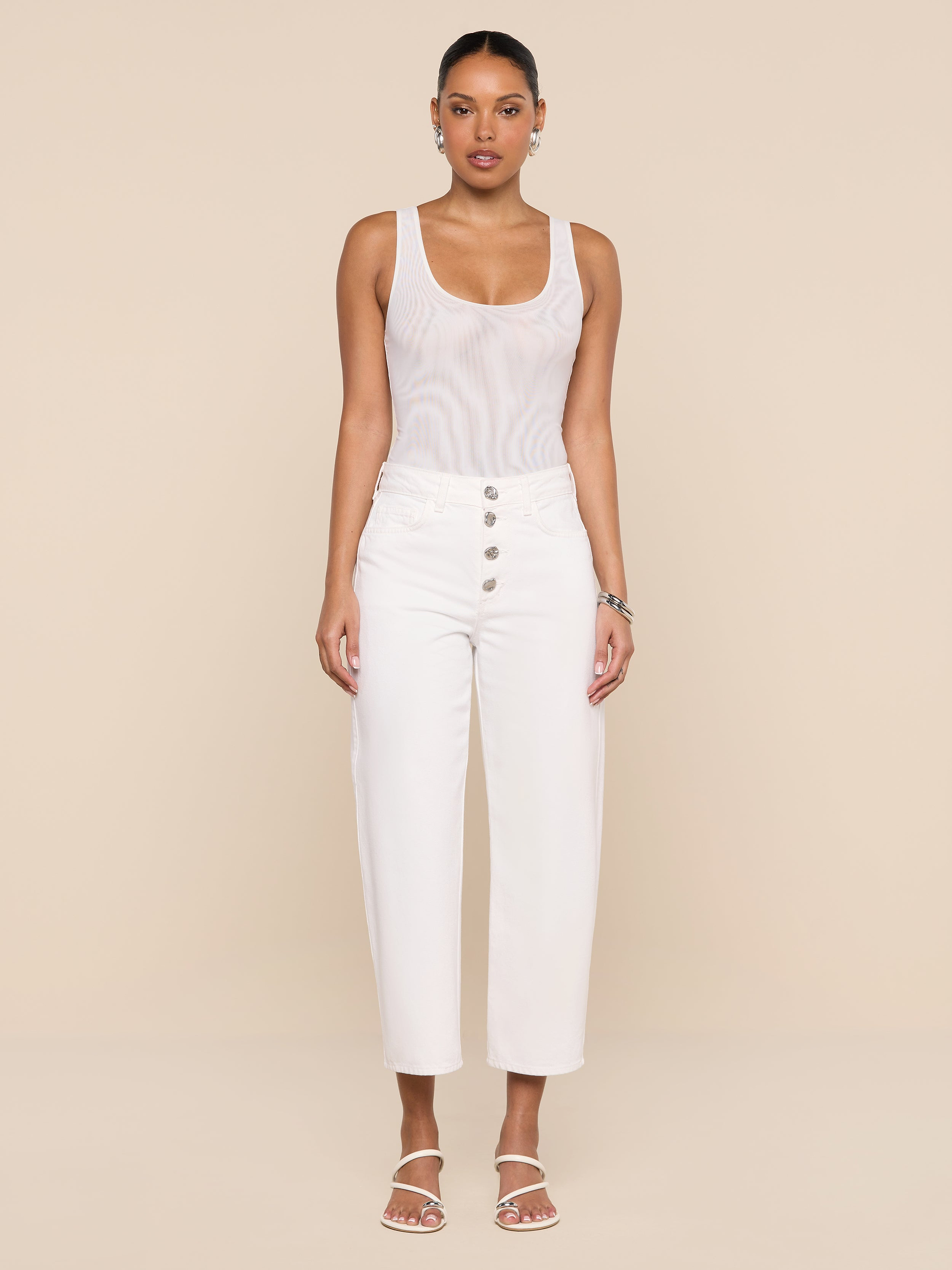 A woman stands facing forward, styled in the Kalea Mesh Bodysuit beneath high-waisted white pants with buttons and white sandals, set against a plain white background.