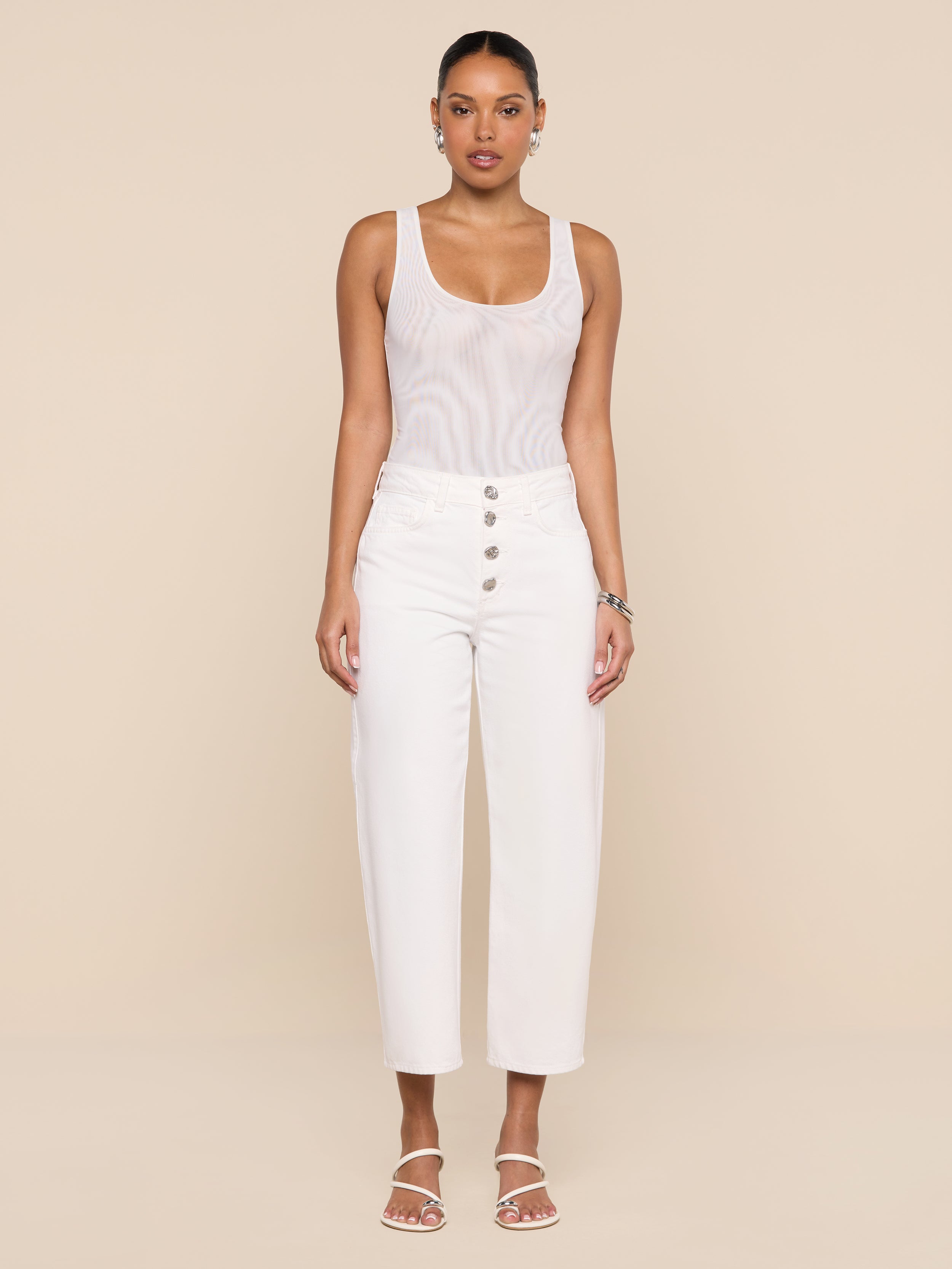A woman stands facing forward, styled in the Kalea Mesh Bodysuit beneath high-waisted white pants with buttons and white sandals, set against a plain white background.