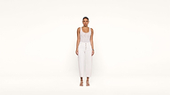 A woman stands against a plain white background, wearing a sleeveless white top, high-waisted white pants with buttons, and white sandals. She looks straight ahead with her arms relaxed at her sides.