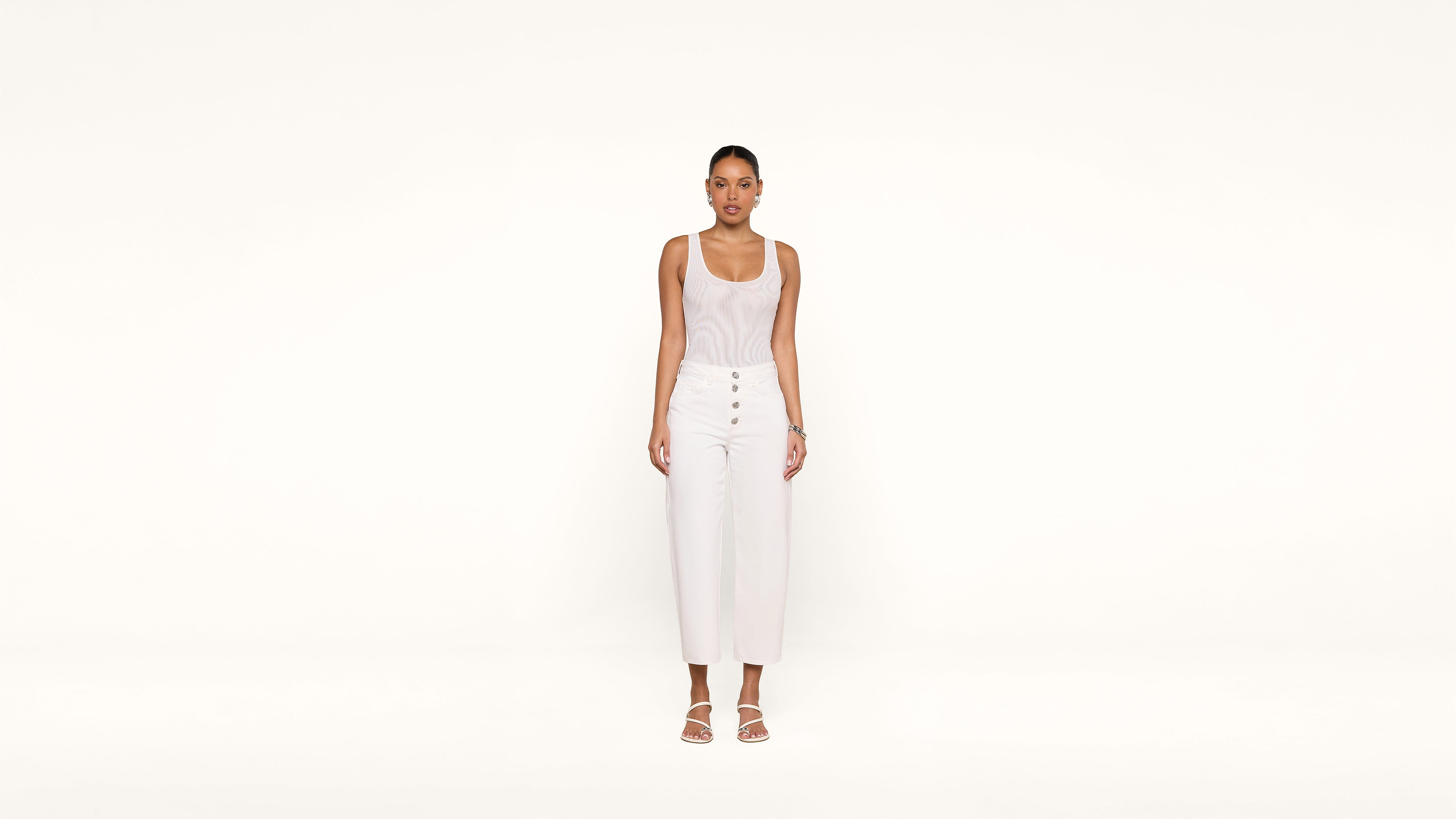 A woman stands against a plain white background, wearing a sleeveless white top, high-waisted white pants with buttons, and white sandals. She looks straight ahead with her arms relaxed at her sides.