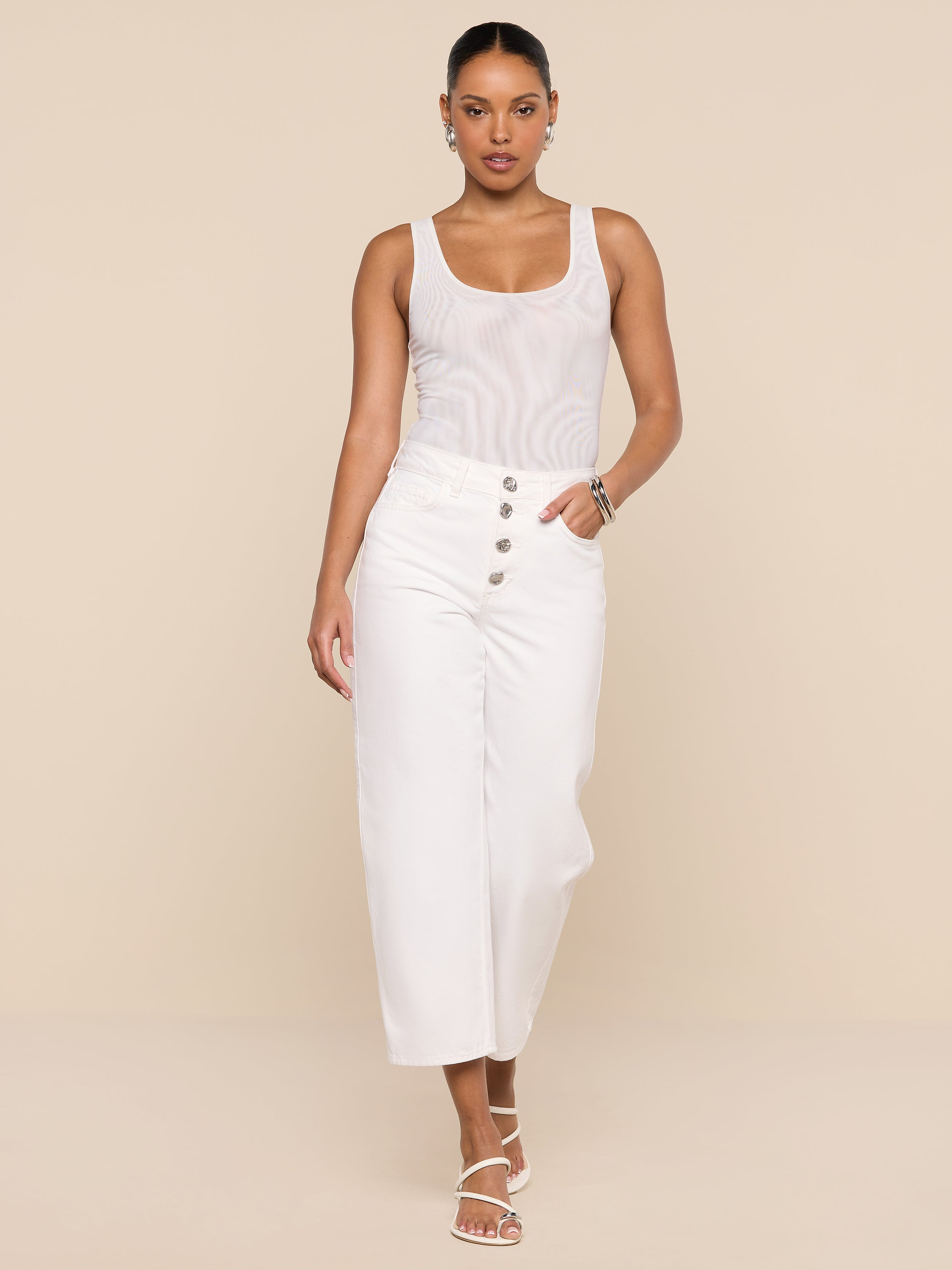 Wearing the Kalea Mesh Bodysuit styled as a white tank top, paired with high-waisted white pants with buttons and white strappy sandals, a woman stands against a plain white background, confidently looking at the camera with one hand in her pocket.