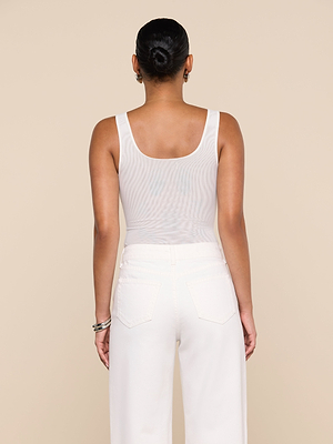 A person stands with their back to the camera, wearing the white Kalea Mesh Bodysuit and white pants. Their hair is in a low bun, with bracelets on their left wrist, against a plain beige background.