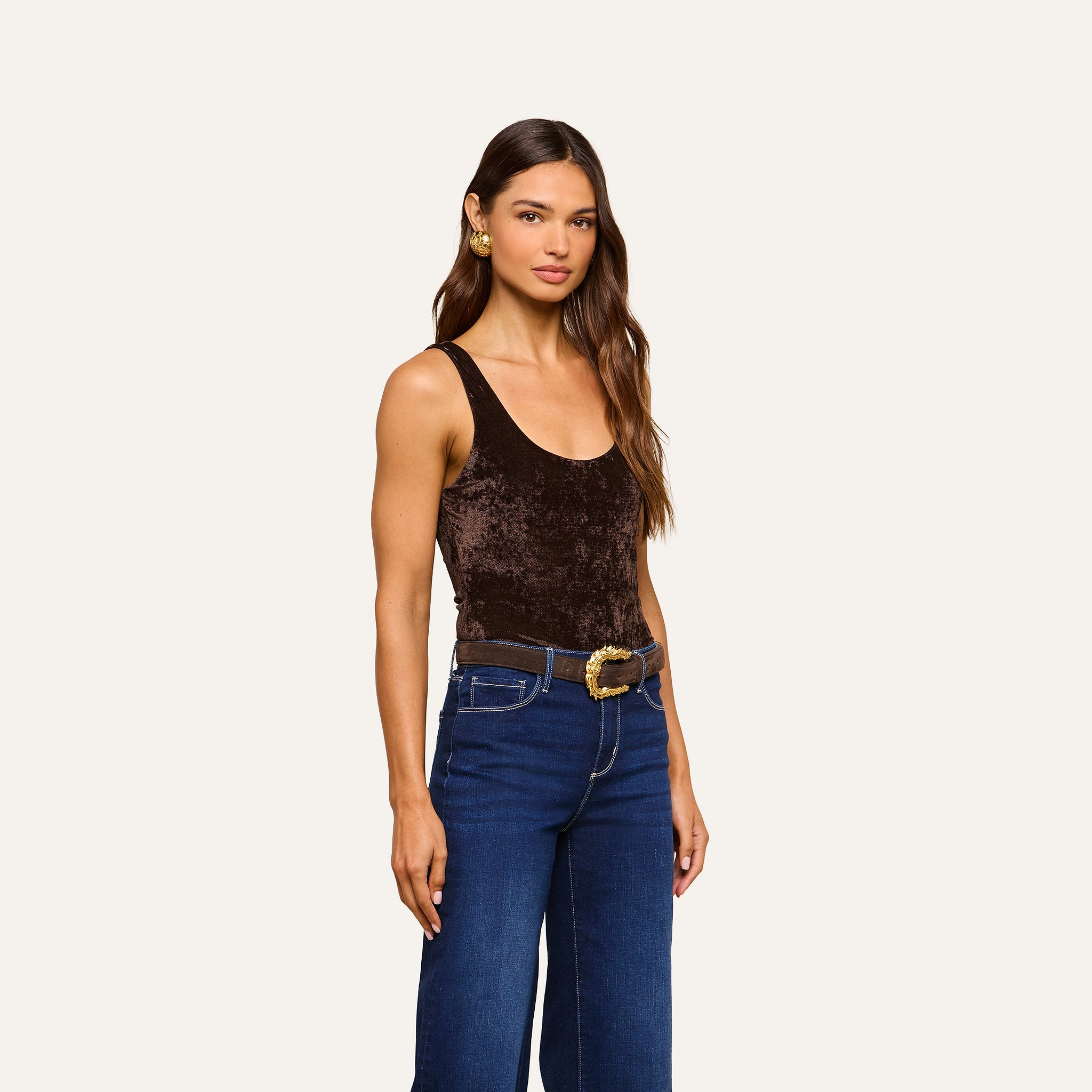 A woman wearing a brown Kalea Velvet Bodysuit, blue jeans, and a wide belt with a decorative buckle stands against a plain light background, looking at the camera.