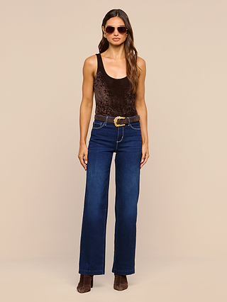 A woman wearing sunglasses, the Kalea Velvet Bodysuit with a scoop neckline, blue high-waisted jeans with a brown belt, and brown boots stands facing forward against a plain beige background.