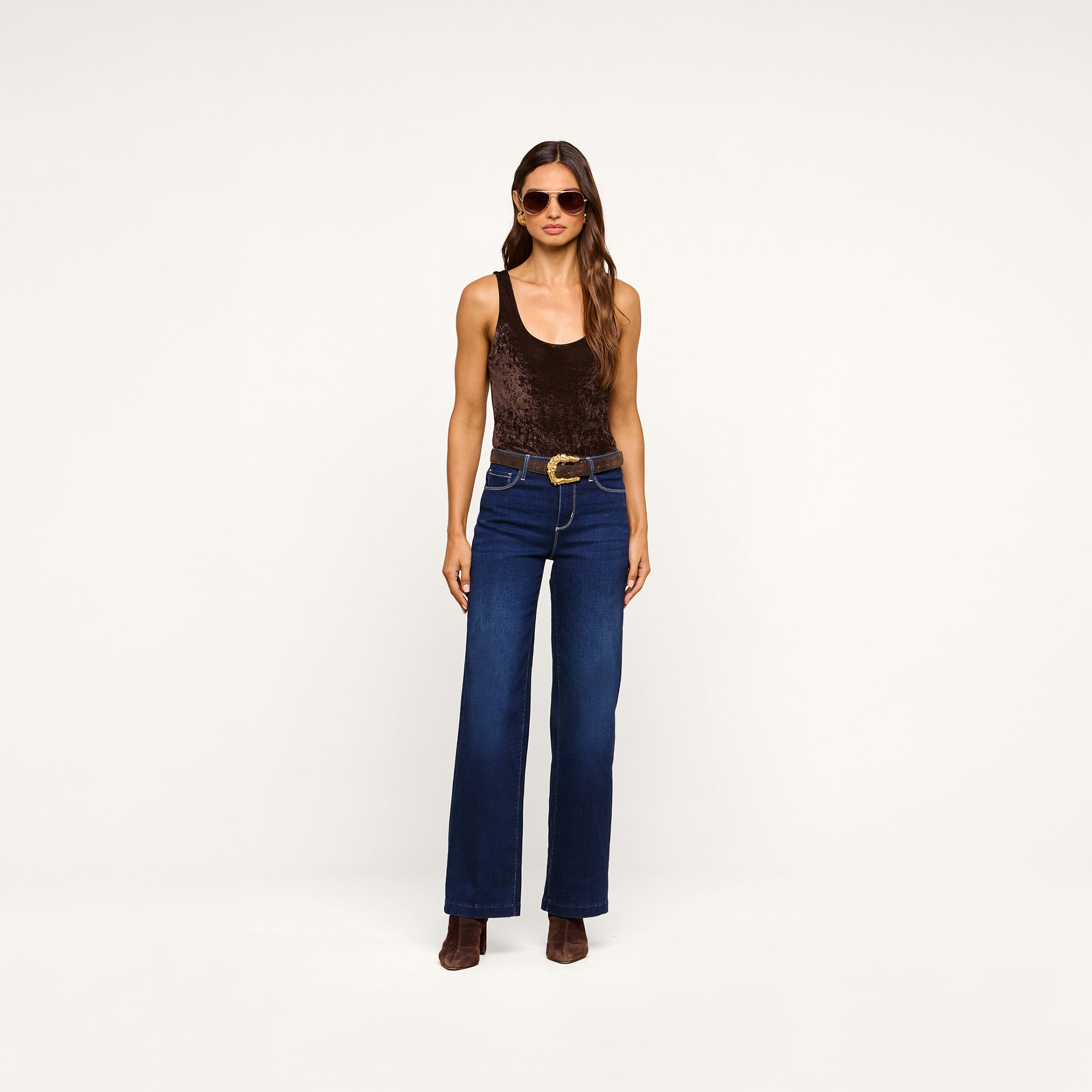 A woman wearing sunglasses, the Kalea Velvet Bodysuit in dark brown, blue high-waisted jeans with a belt, and brown boots stands against a plain white background.