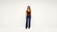 A woman wearing sunglasses, a brown sleeveless top, blue high-waisted wide-leg jeans, a belt, and brown boots stands facing the camera against a plain white background.