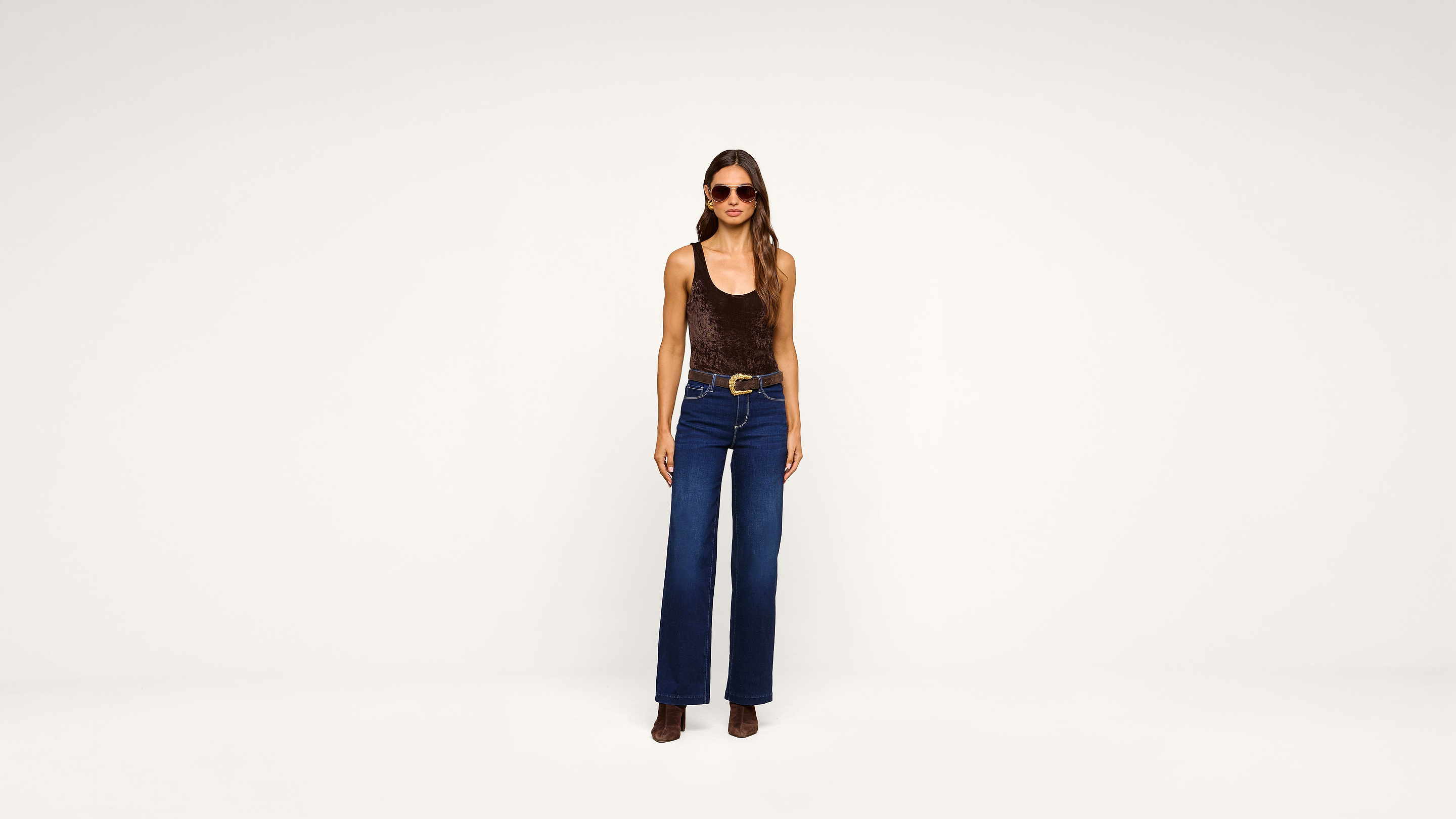 A woman wearing sunglasses, a brown sleeveless top, blue high-waisted wide-leg jeans, a belt, and brown boots stands facing the camera against a plain white background.