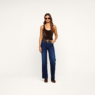 Woman wearing sunglasses, the Kalea Velvet Bodysuit layered with a lightweight outer piece, blue high-waisted wide-leg jeans with a belt, and brown boots stands against a plain white background.