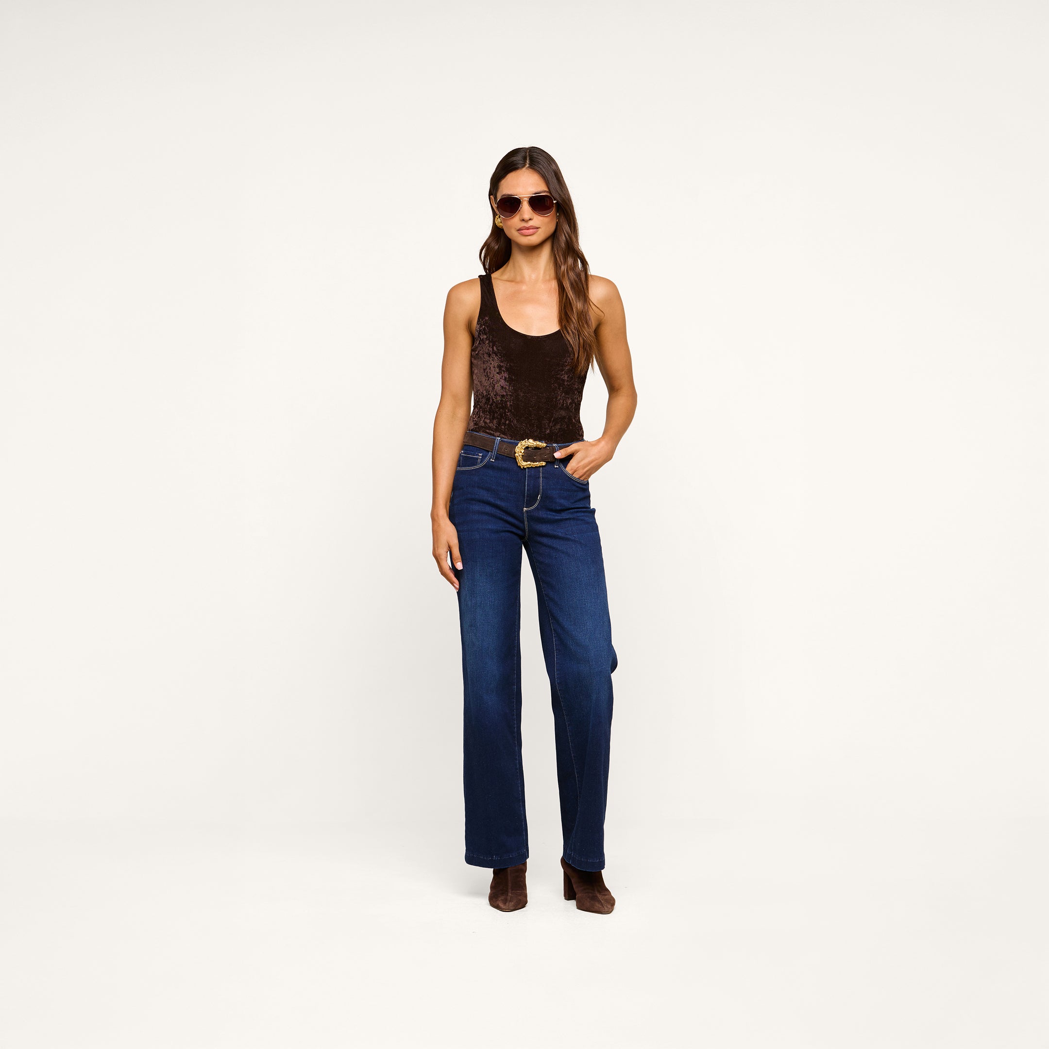 Woman wearing sunglasses, the Kalea Velvet Bodysuit layered with a lightweight outer piece, blue high-waisted wide-leg jeans with a belt, and brown boots stands against a plain white background.