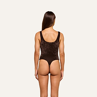 A woman with long brown hair, seen from behind, is wearing the Kalea Velvet Bodysuit—a sleeveless dark brown velvet bodysuit with a thong back—standing against a plain light background.