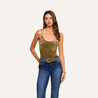 A woman with long brown hair wears the Kalea Velvet Bodysuit, which features a flattering scoop neckline in a luxurious gold velvet. She pairs it with blue jeans, a wide belt, and a bracelet, standing against a beige background and gazing at the camera with one hand resting by her side.