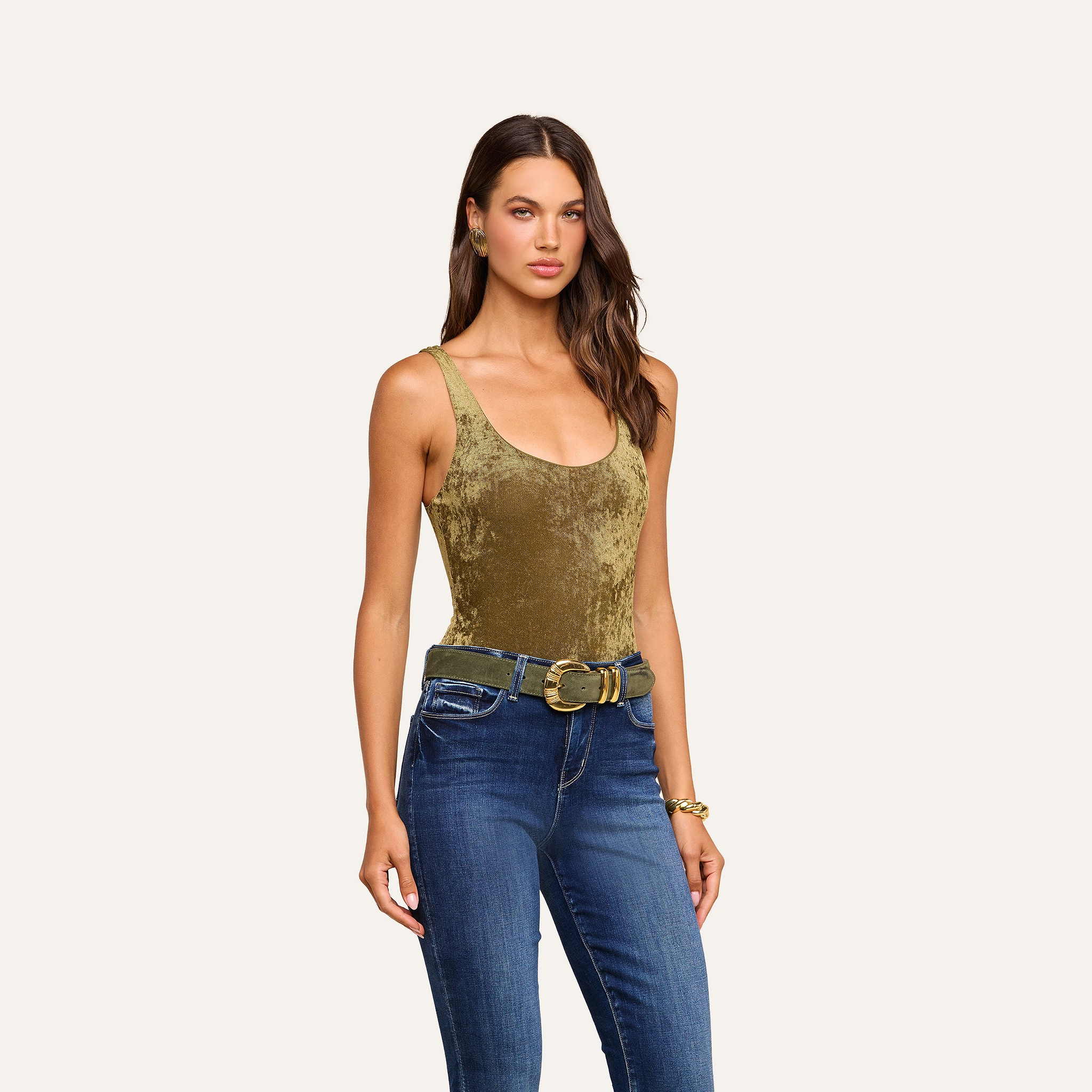 A woman wearing the Kalea Velvet Bodysuit in olive green, styled with blue jeans, a wide green belt, gold earrings, and a gold bracelet stands against a beige background.