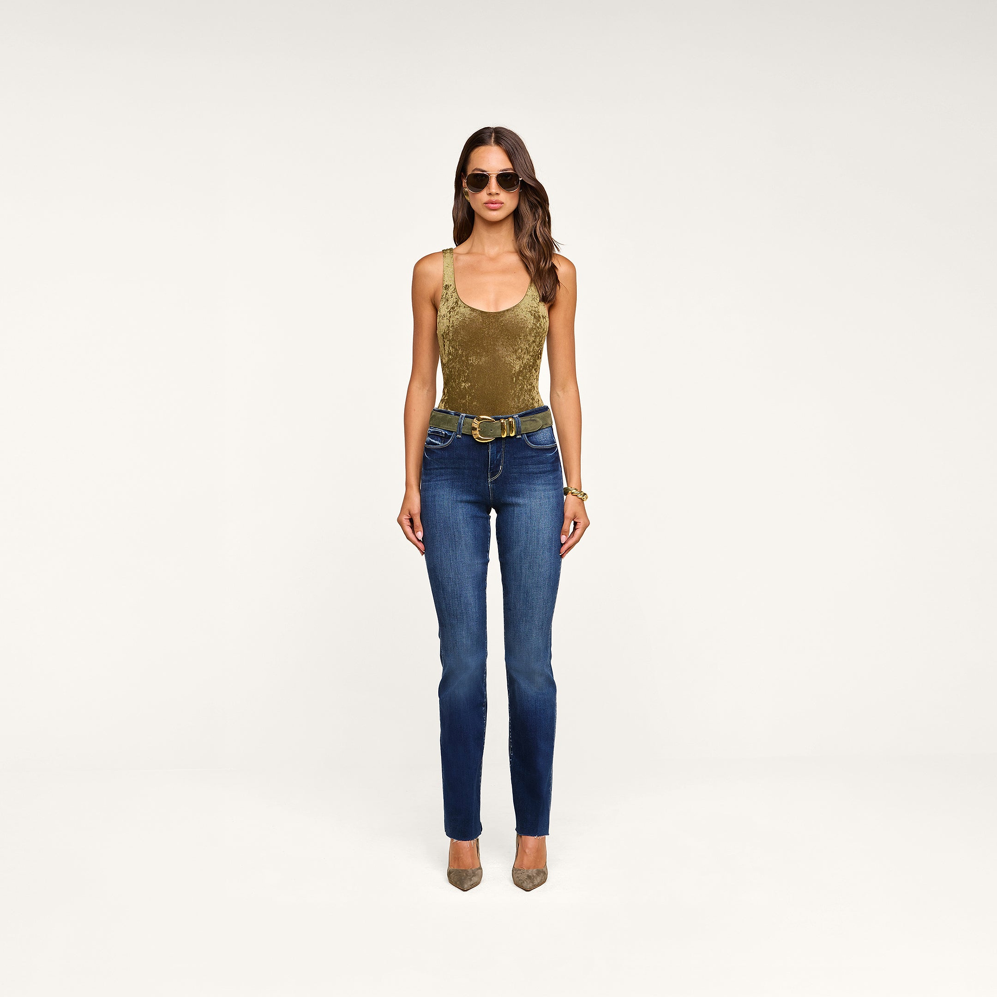 A woman stands against a beige background, wearing sunglasses, layered bracelets, tan heels, blue jeans with a double-buckle belt, and the Kalea Velvet Bodysuit in olive green featuring a stylish scoop neckline.