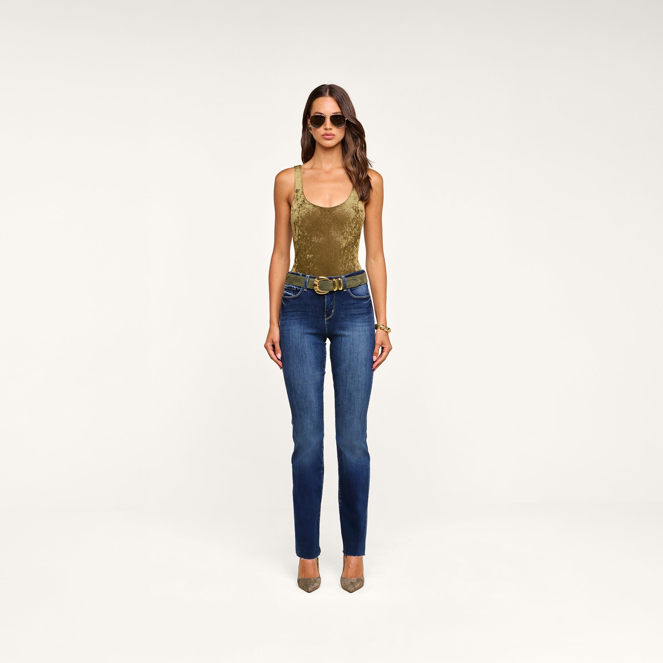 A woman stands against a beige background, wearing sunglasses, layered bracelets, tan heels, blue jeans with a double-buckle belt, and the Kalea Velvet Bodysuit in olive green featuring a stylish scoop neckline.
