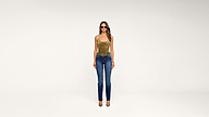 A woman wearing sunglasses, a gold tank top, blue jeans, and heels stands against a plain white background.