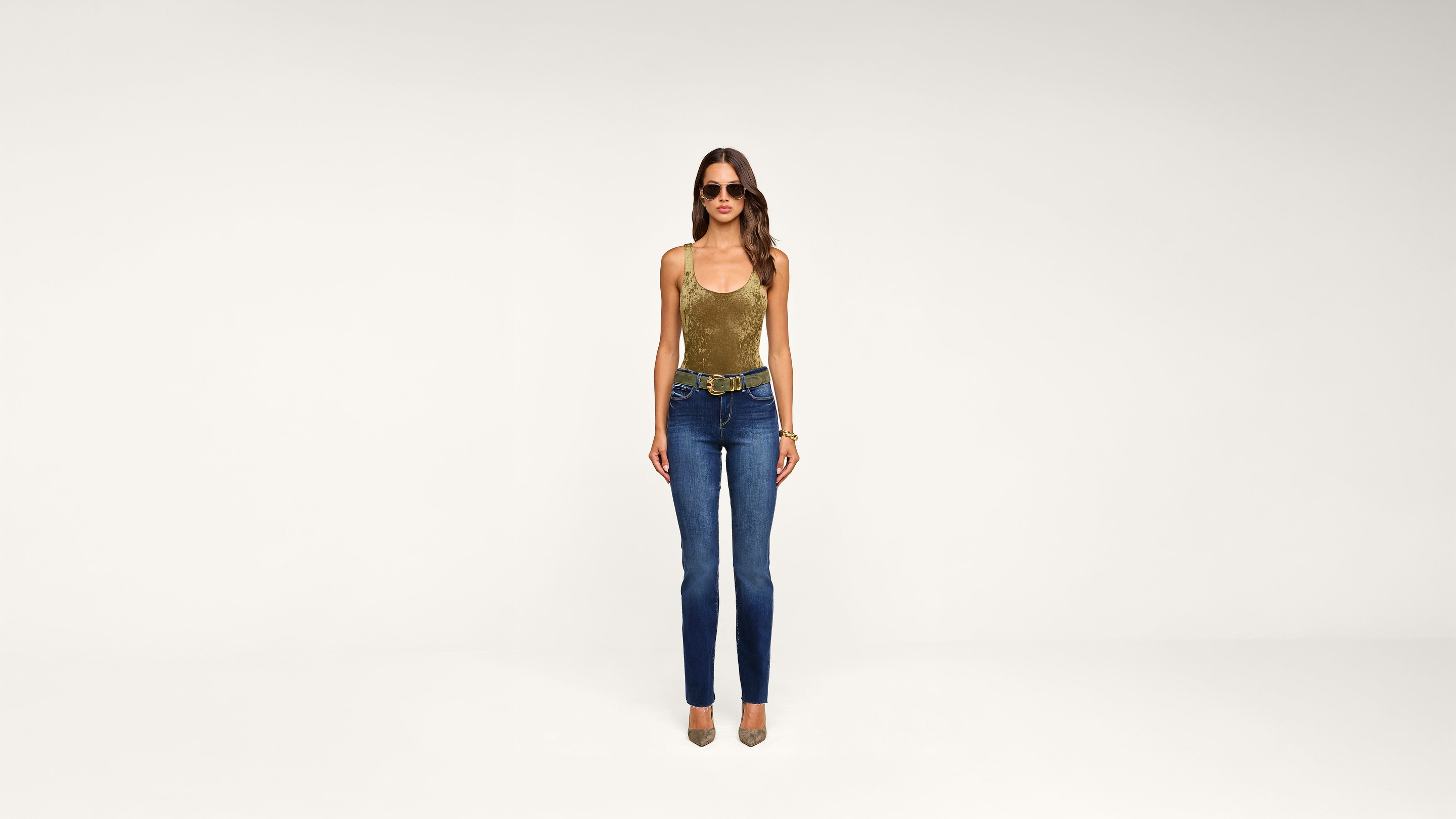 A woman wearing sunglasses, a gold tank top, blue jeans, and heels stands against a plain white background.