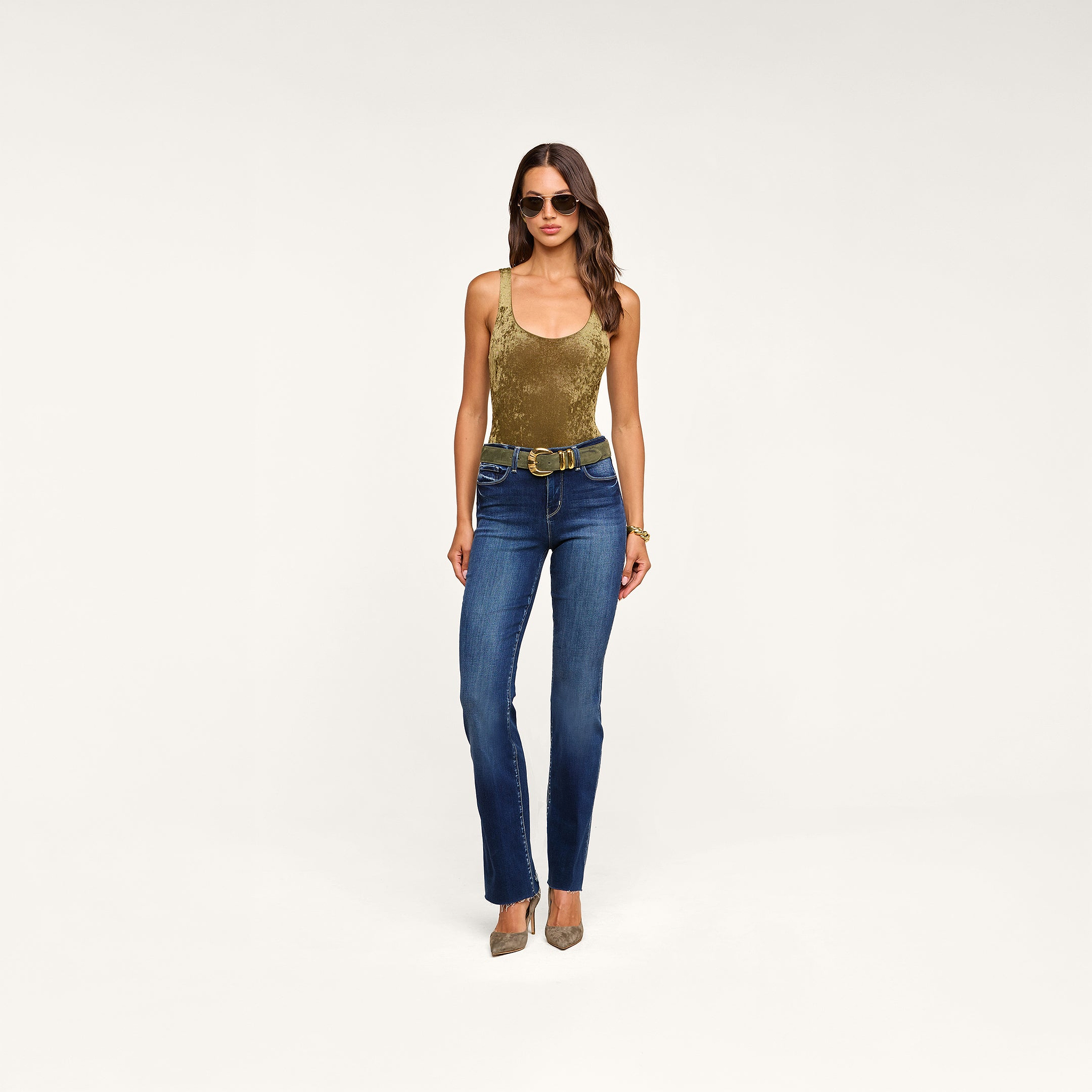 A woman wearing sunglasses and the Kalea Velvet Bodysuit—a sleeveless gold velvet piece with a scoop neckline—paired with belted blue jeans and taupe high heels, stands against a plain beige background.