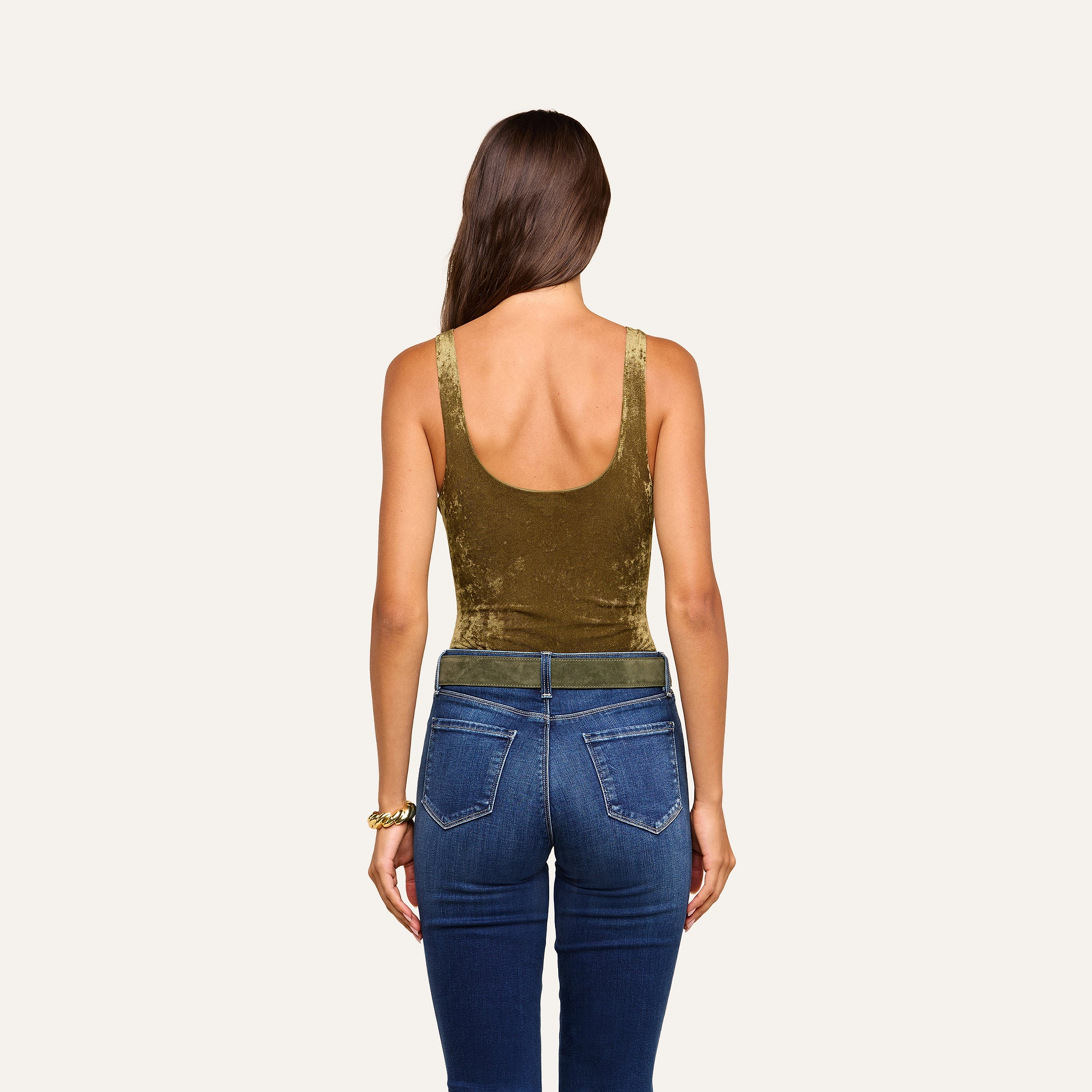A woman with long brown hair, wearing the Kalea Velvet Bodysuit in olive green—which features a sleeveless design and scoop neckline—paired with blue jeans, stands facing away from the camera against a plain beige background.