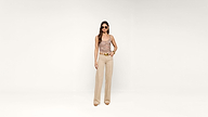 A woman wearing sunglasses, a patterned sleeveless top, beige wide-leg pants, and heels stands against a plain white background.