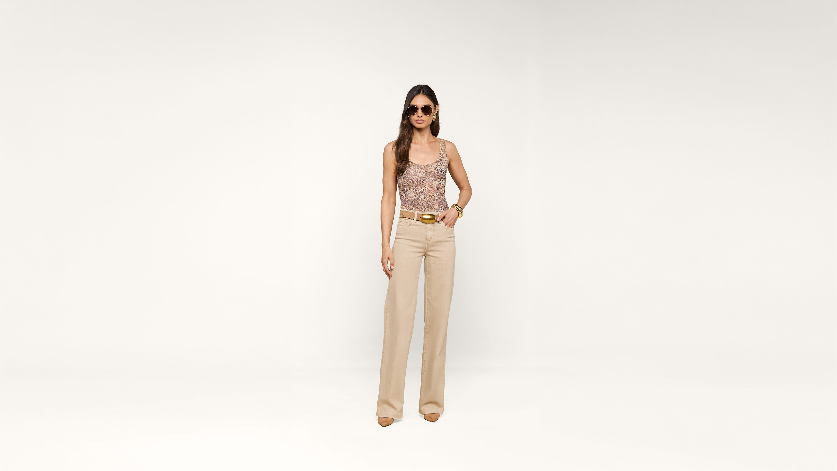 A woman wearing sunglasses, a patterned sleeveless top, beige wide-leg pants, and heels stands against a plain white background.