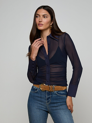 A woman with long brown hair wears the Isola Mesh Blouse over a dark top, styled with blue jeans, a tan belt with a gold buckle, and gold hoop earrings. She stands against a light gray background, looking to the side.