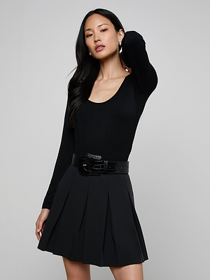 A woman with long dark hair poses against a gray backdrop, wearing the Cassandra Ruched Bodysuit—a fitted black long sleeve top with a scoop neckline—styled with a black pleated skirt and wide belt, one hand behind her head.