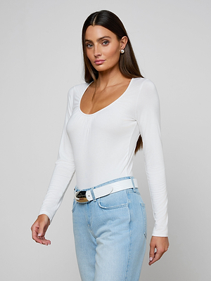A woman with long dark hair wears the Cassandra Ruched Bodysuit, a fitted white long sleeve top with a scoop neckline, paired with light blue high-waisted jeans and a white belt, standing against a plain light background.