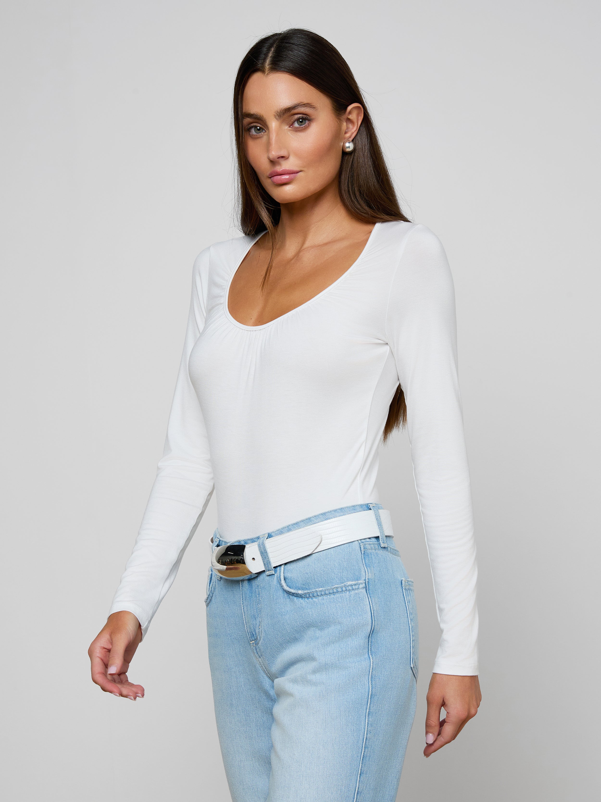 Woman with long brown hair wearing the Cassandra Ruched Bodysuit—a white long sleeve top with a scoop neckline—paired with light blue jeans and a white belt, standing against a plain light background.