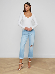 The woman is wearing the Cassandra Ruched Bodysuit, a white long-sleeve top with a scoop neckline, paired with light blue ripped jeans and high-heeled sandals. She stands against a plain light background, looking to the side with her hands resting on her jeans.