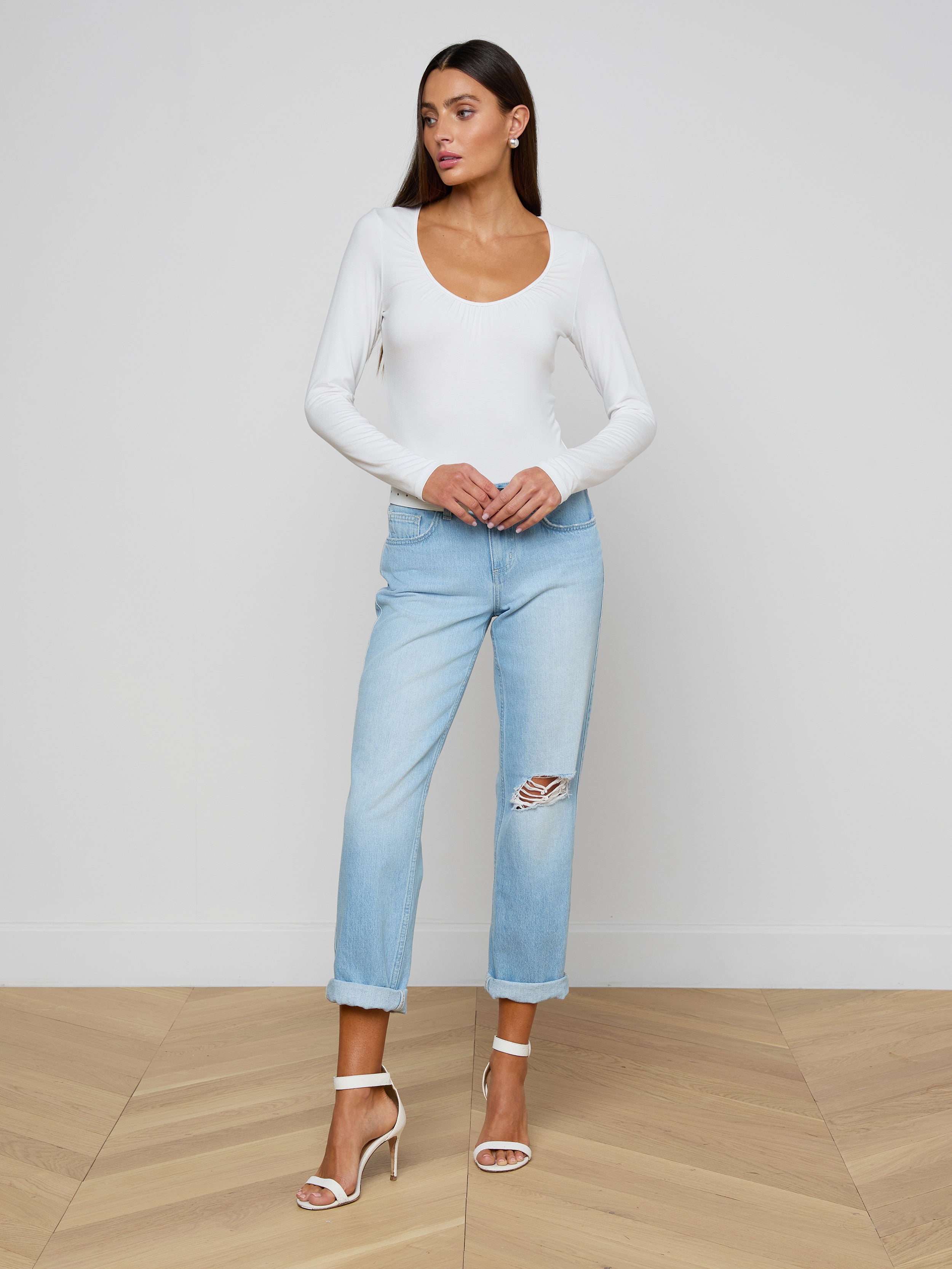 The woman is wearing the Cassandra Ruched Bodysuit, a white long-sleeve top with a scoop neckline, paired with light blue ripped jeans and high-heeled sandals. She stands against a plain light background, looking to the side with her hands resting on her jeans.